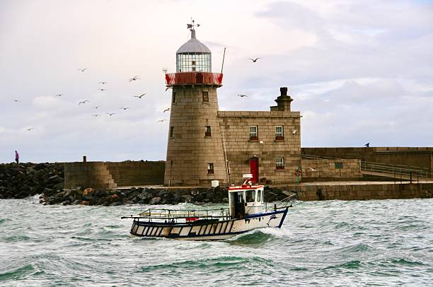 view of Hook Lighthouse, located on Hook Head in County Wexford, Ireland. The lighthouse stands tall against a dramatic sky with moody clouds. The foreground features a rocky coastline, with layers of textured rocks leading to the ocean.