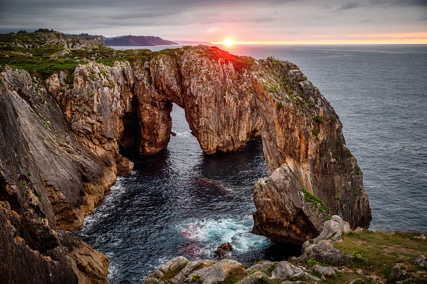 Glowing orange and red hues of the sunset illuminating the cliffs of Guadamia in Pria, Asturias, create a stunning natural arch rising majestically over the tranquil sea