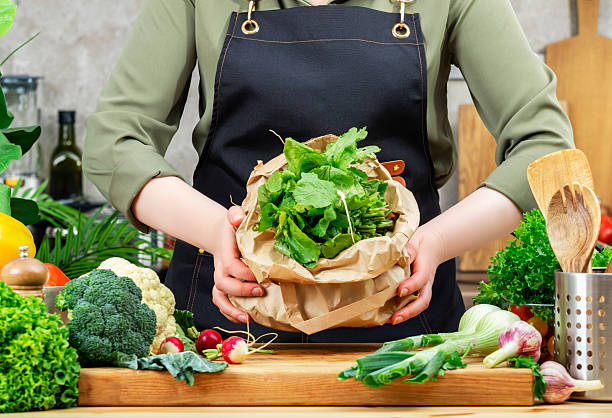 istock Chef holds vegetables peels in paper bag for processing compost. Wooden table, kitchenware, vegetables and herbs. Healthy eating, zero waste, blogging, recycling 2218498661