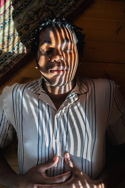Sunlight filtering through window blinds casting striped shadows across young person relaxing on wooden floor, embodying tranquil introspective moment