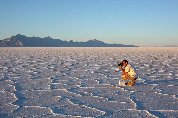 Individual in vibrant red jacket crouches on expansive salt flats under dramatic sunset sky to take a picture. Stunning nature landscape with reflective water surface. Twilight solitude and reflection.