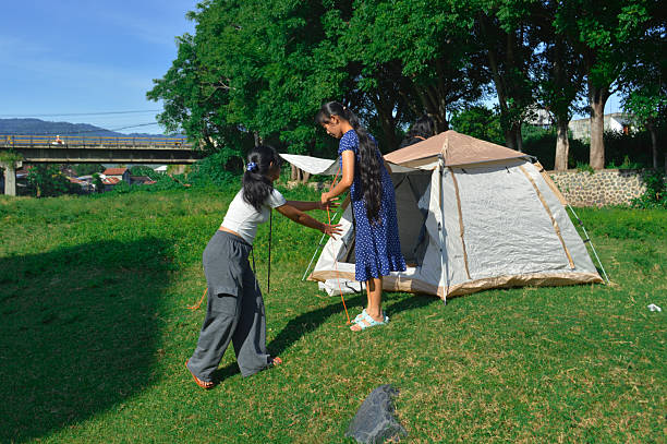 Two girls hold and stretch the canopy ropes of the camping tent while adjusting its setup in the grassy field.