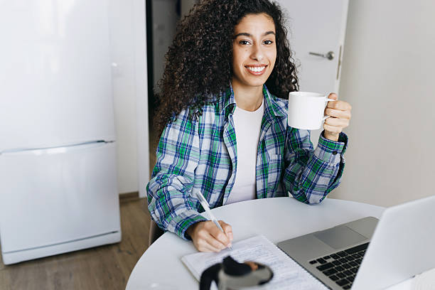 istock Female student with dark skin and curly hair doing homework at kitchen table using laptop, making brief summary of video tutorial, writing in her copybook, holding mug of coffee and smiling at camera 2218539207