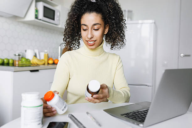 istock Black girl restocking B-vitamins or poly-unsaturated fatty acids, making order online on marketplace holding bottles of biologically active supplements sitting at kitchen table in front of laptop 2218539362
