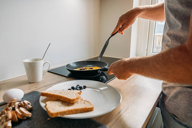 Close-up of a man cracking an egg into a pan on a wooden counter. Sliced bread and mushrooms on a cutting board. Cozy morning routine with coffee mug in kitchen