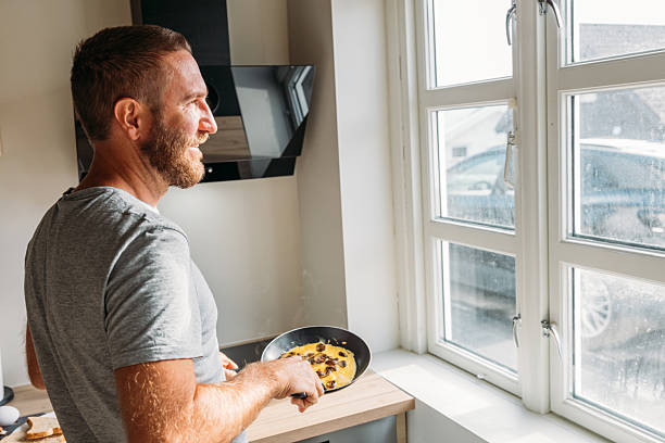 Man smiling while holding a frying pan with omelette by a window. Bright kitchen scene with natural light. Casual breakfast preparation in home setting.