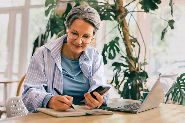 istock Mature female freelancer taking notes and using phone, working remotely in relaxing coffee shop 2218552861