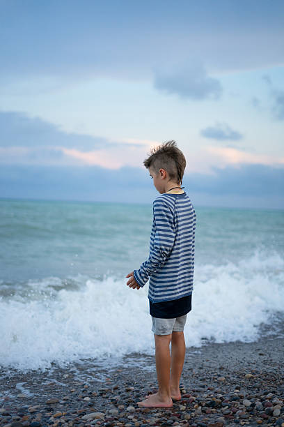 Young boy stands barefoot on a rocky beach, watching the waves crash in the evening