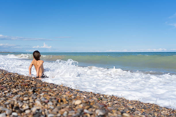 Child sitting on the shore of a pebble beach is playing with the waves of the sea during summer vacation