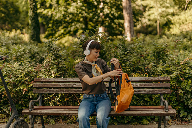 istock Young stylish woman searching something in her backpack while sitting on a bench in a park 2218578846