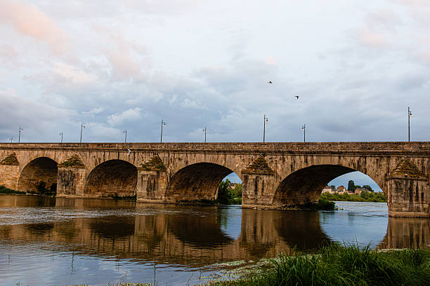 A historic stone bridge with multiple arches spans a calm river, reflecting in the water. The sky is partly cloudy with a hint of sunset colors, and two birds are flying above.