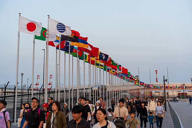 Osaka, Japan - June 1 2025 : Crowds leaving Expo 2025 Osaka, Kansai, Japan under fluttering international flags at the East Gate during a vibrant dusk sunset.