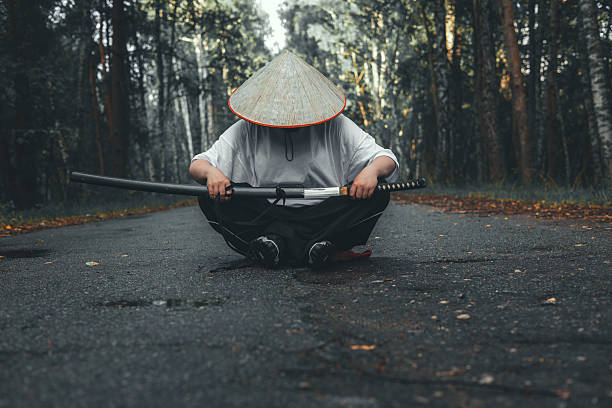 A warrior in a straw hat sits on a path in the forest, holding a katana in both hands. A female warrior practices meditation before a fight. A warrior sits on the road and unsheathes a katana..