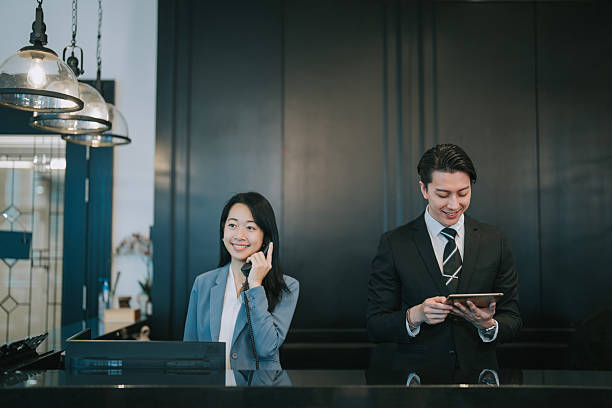 Asian hotel concierge team at the reception counter—female staff answering a phone call while her male colleague uses a digital tablet. Depicts professional hospitality service in a sleek, modern hotel setting. Ideal for themes related to customer service, digital check-in, and business travel.hotel receptionist, front desk, concierge service, hotel staff, Asian professionals, business attire, modern hotel, hospitality industry, customer service, digital check-in, smiling receptionist, teamwork, luxury hotel, business travel, front desk counter, lobby area, corporate hospitality, digital tablet, technology in hospitality, welcoming service,