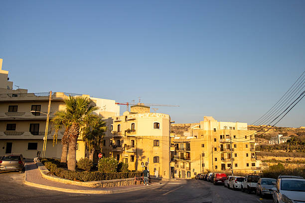 People on Triq Ta' Barda, a street located in the coastal village of Marsalforn, situated on the island of Gozo, Malta. Marsalforn is known for its scenic bay, making it a popular destination for both locals and tourists. Logos and insignia are visible.