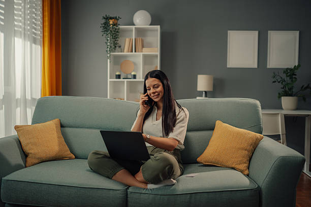 istock Young woman talking on phone and using laptop on sofa at home 2218611511