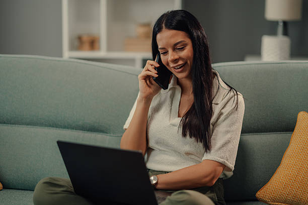 Young woman working from home and talking on the phone while looking at laptop