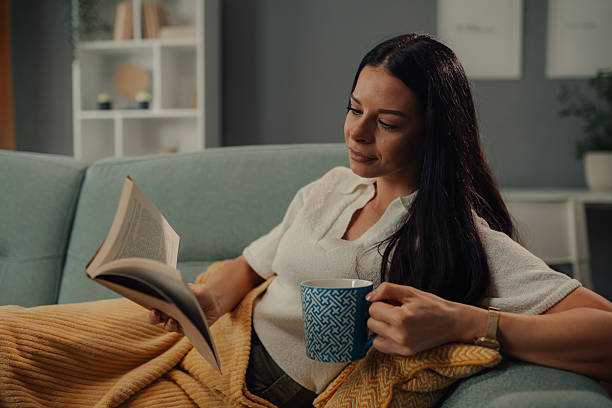 istock Young woman relaxing on sofa reading a book and drinking tea 2218612104