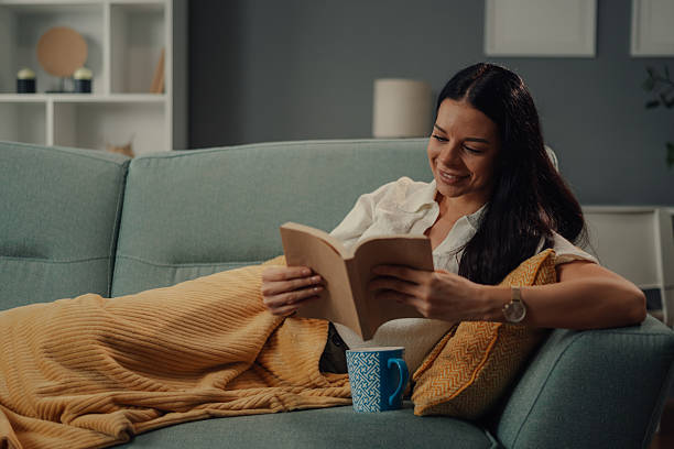 istock Young woman relaxing on sofa reading a book and drinking tea 2218612116