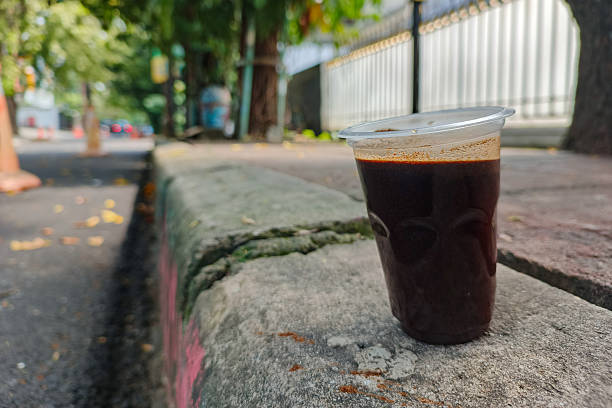 A plastic cup of black coffee sits on a sidewalk ledge beside a quiet street.