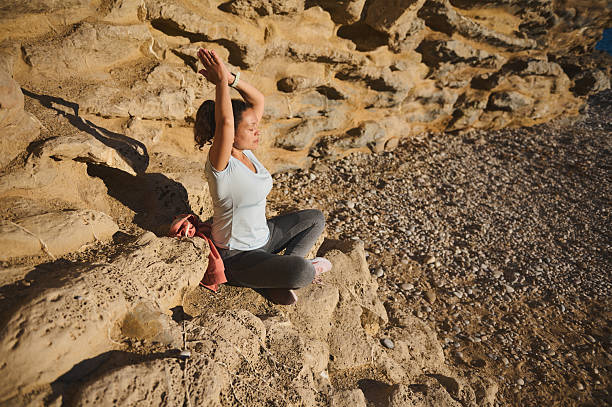 A serene moment captured of a woman engaging in meditative yoga poses, set against a rugged coastal rock background bathed in sunlight.