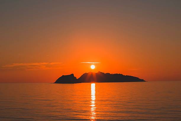 A stunning sunset over a calm sea with an island silhouette under a vibrant orange sky.