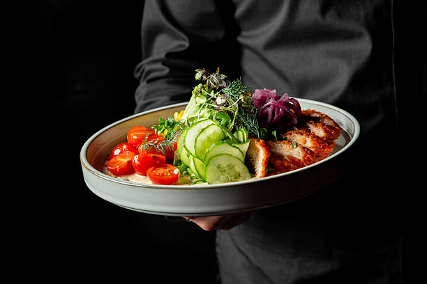 A chef presents a gourmet salad with fresh ingredients including cherry tomatoes, cucumbers, mixed greens, pickled onions, and grilled chicken, beautifully plated and ready to serve