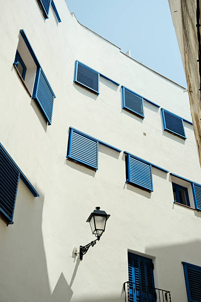 Displaying a white facade of a mediterranean-style building, featuring blue shutters under the bright sunlight of a clear day
