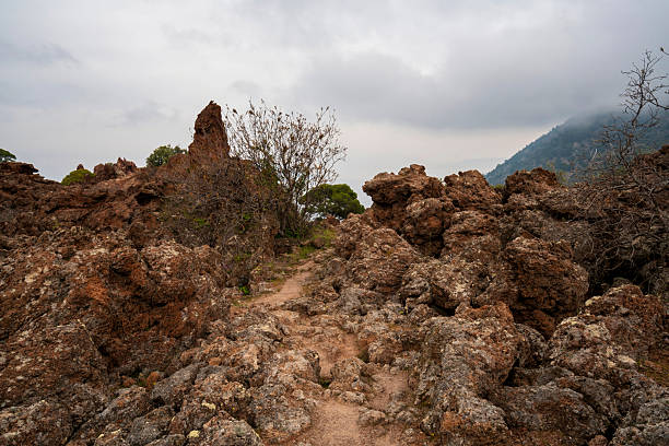 A narrow dirt path winds through rugged lava rock formations and dry brush on the volcanic slopes of Kameno Vouno in Methana.