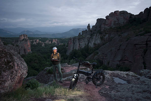 Sustainable travel & outdoor nature lifestyle. Cyclist with e-bike on rocky setting, illuminated by device under blue hour sky. Adventure & technology. GPS tracking.