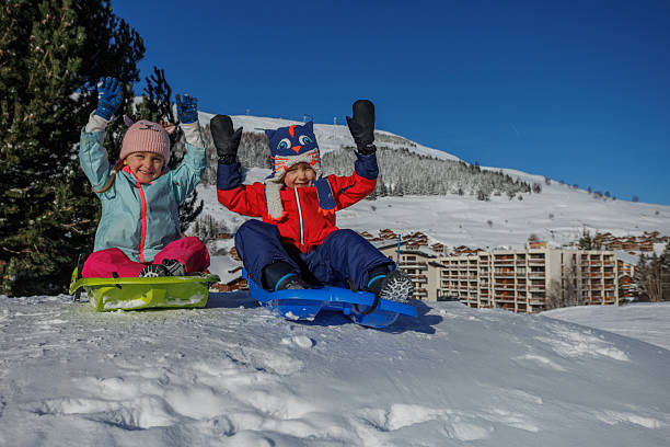 Two children ride a green sled with chalet views in background
