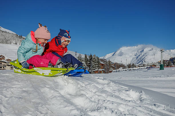 istock Two kids sled down a snowy hill, bundled up and smiling 2218683980