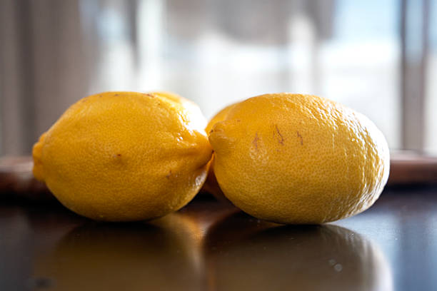 Two fresh lemons resting on a reflective dark surface in natural light, highlighting their vibrant color and textured skin.