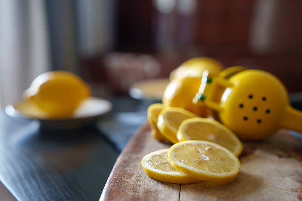 Wooden cutting board with fresh lemon slices and a yellow handheld citrus juicer placed nearby, with whole lemons in the background on ceramic plates.