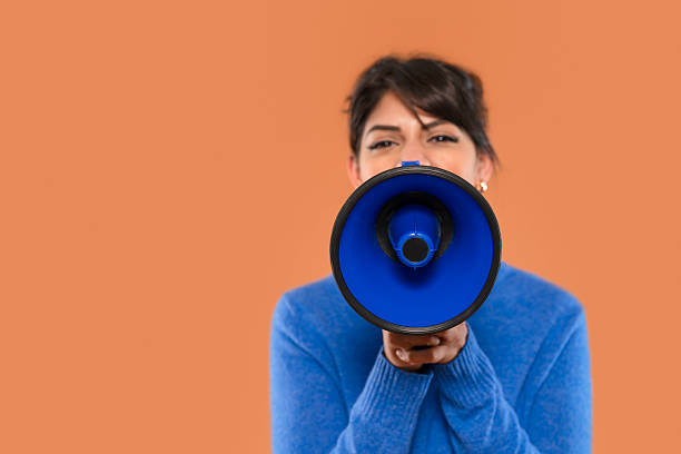 istock Woman in blue sweater holds a blue megaphone while speaking, emphasizing her message in front of brown backdrop 2218696458