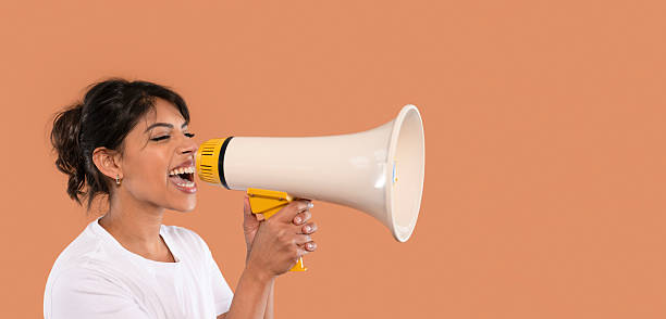 istock Woman enthusiastically using a megaphone to communicate a message in a minimalist setting with a warm, solid colored background 2218696648