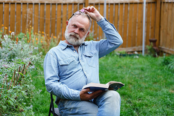 Man sits on a chair in a garden, reading a book. He adjusts his glasses, surrounded by vibrant flowers and grass, enjoying a tranquil afternoon outdoors.