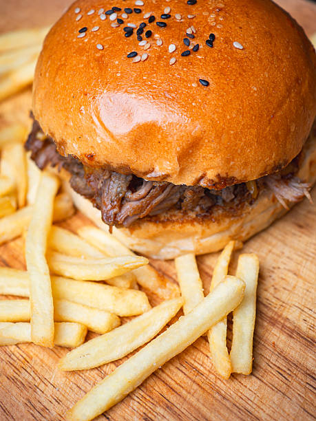 Pulled pork hamburger with sesame bun served with crispy French fries on rustic wooden surface.