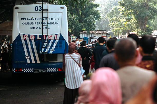 People queue behind an Indonesian police mobile license renewal bus parked under trees.