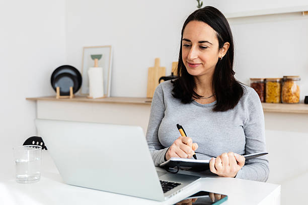 istock Woman working remotely from home kitchen taking notes from laptop 2218713879