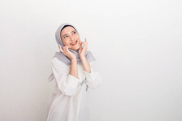 cheerful woman wearing a hijab smiling brightly, with her hands near her face, evoking feelings of happiness, confidence, and contentment. The light background emphasizes her joy and serene expression