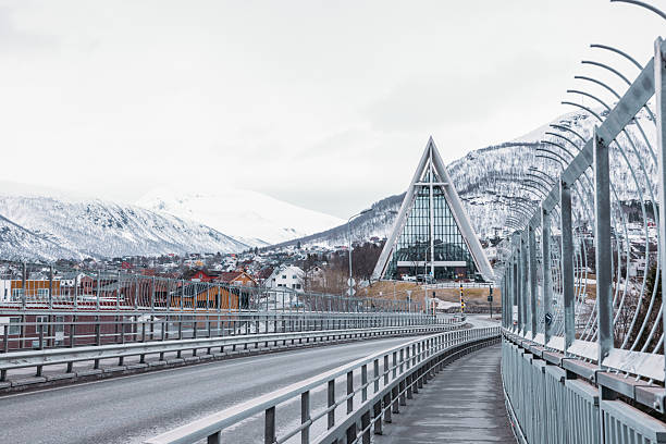 Icy road leading to Tromsø's Arctic Cathedral, framed by snowy peaks. Scenic winter view of modern architecture. Iconic landmark in Nordic town.