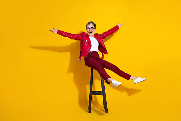 istock Cheerful elderly stylish woman in red leather jacket posed enthusiastically on a bright yellow background sitting on a stool 2218721247