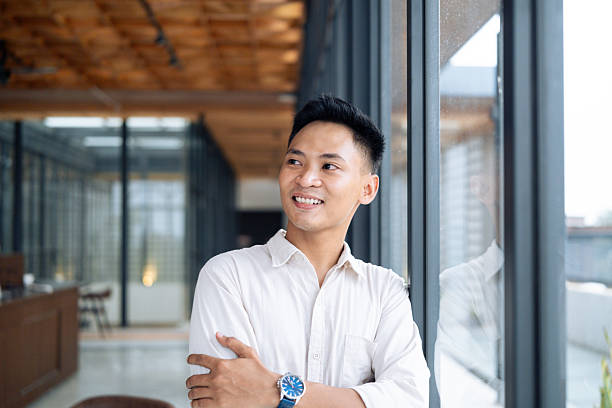 istock Confident young man in a white shirt smiling and leaning casually by large windows in a modern office setting, embodying positivity and professionalism. Bright natural lighting enhances the workplace 2218722278