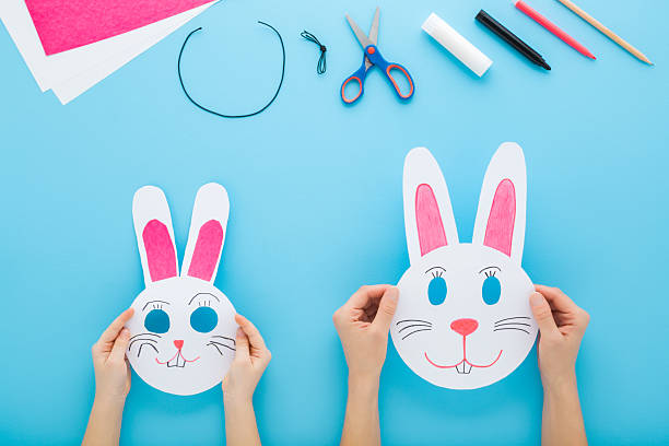 istock Mother and child hands together making white smiling rabbit paper masks on light blue table background. Pastel color. Point of view shot. Closeup. Creating easter decoration. Top down view. 2218722634