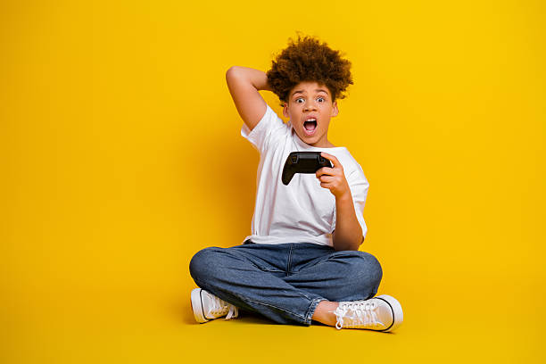 istock Excited boy holding a game controller sitting on a vibrant yellow background showing surprise and enthusiasm 2218722787