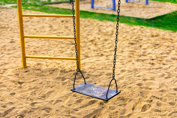 istock Worn black swing hanging from chains over a sandy playground with climbing frames in the background. 2218724233