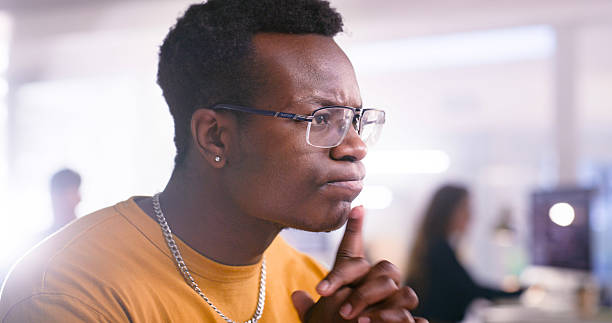 A young professional is deeply focused and contemplating ideas in a modern workplace. The image conveys thoughtfulness and productivity captured with soft lighting and a technologically centered environment.