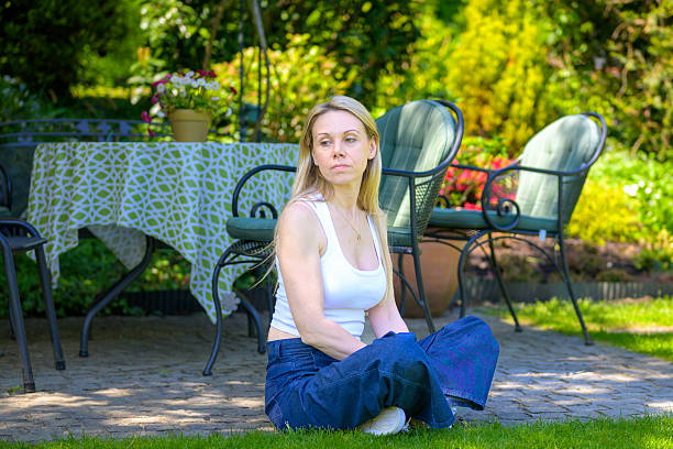 A blonde woman sits cross-legged on the edge of a garden patio, looking thoughtfully into the distance.