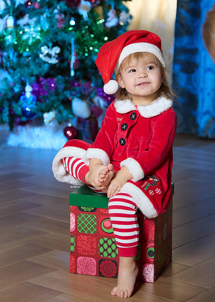 Cheerful little girl in Santa's helper costume is sitting on the box of the Christmas gift with leg on leg. Shooting at a home